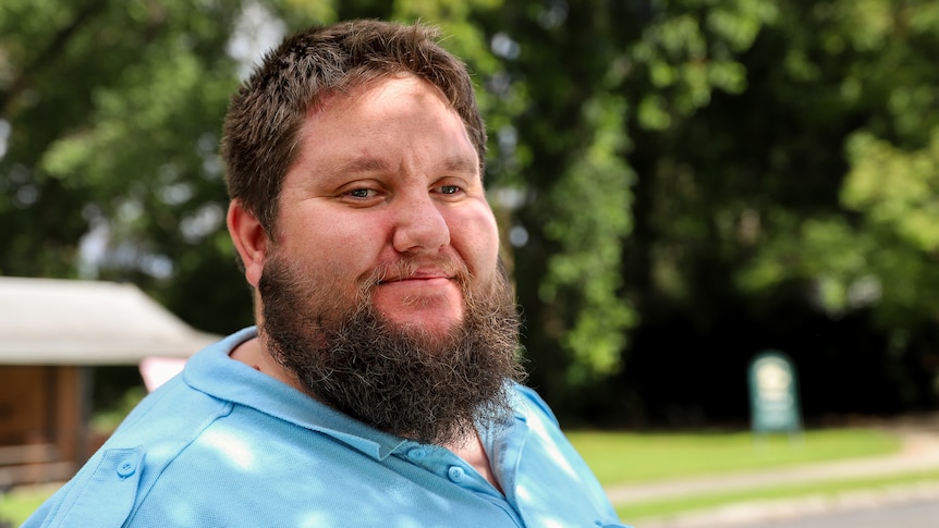 Man with beard smiles in dappled light wearing taxi driver uniform with green trees behind in country street