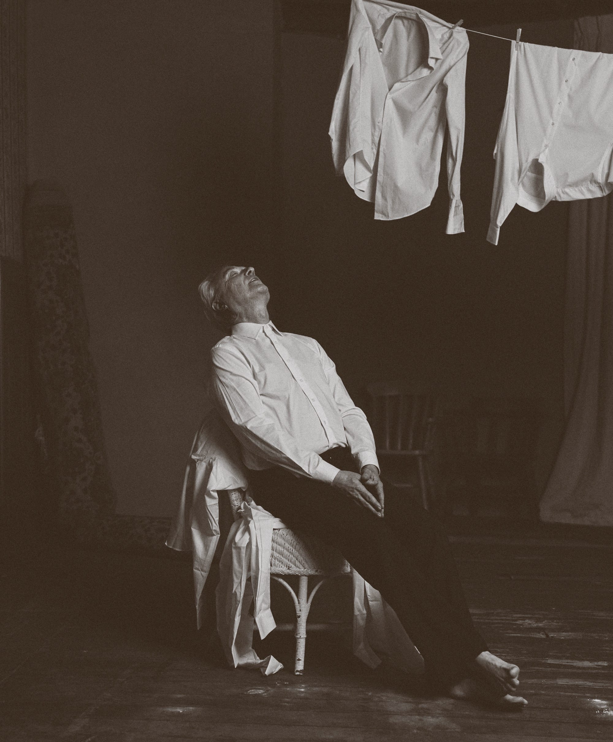 a black and white photo by Austn Fischer of david byrne sitting on a chair beneath hanging shirts
