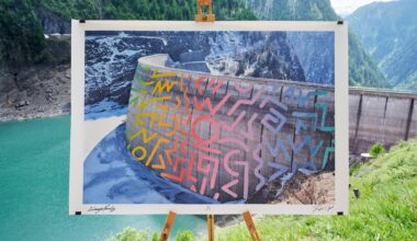 A photographic print of a dam in Switzerland with a street art intervention superimposed on the image, which is photographed on an easel in front of the same landscape