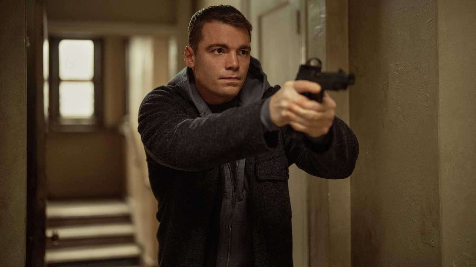 Gabriel Basso as Peter Sutherland pointing a gun while in a hallway in The Night Agent season 3