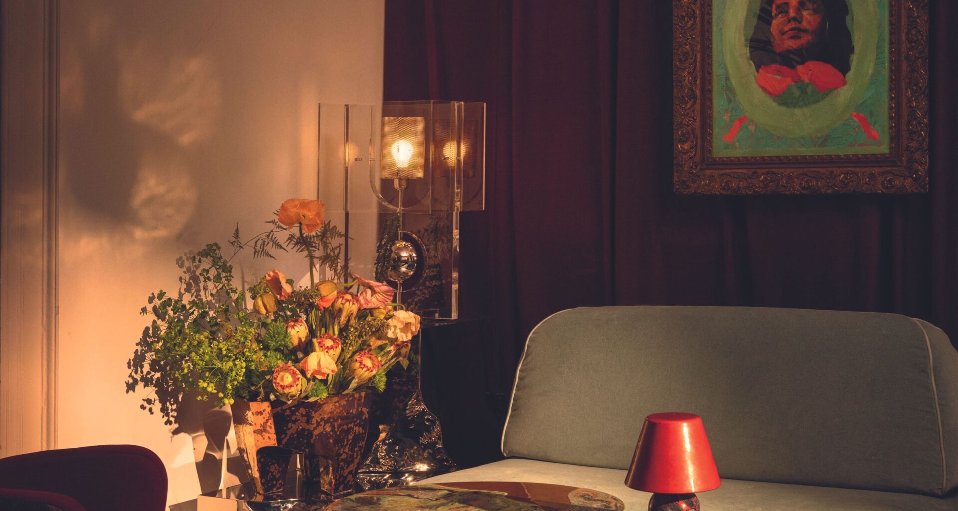 A sun-lit living room decorated with artisanal pieces in glass, wood, marbled copper, fresh flowers, plush, velvety seats, and a colorful artwork hanging on the wall.
