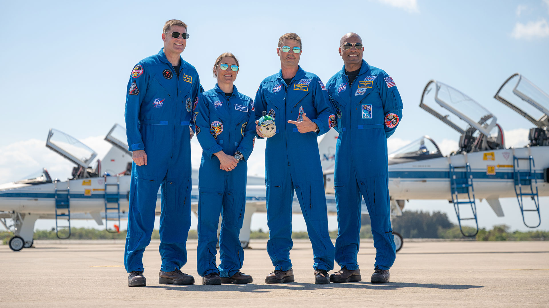 three men and a woman, each wearing a blue flight suit, pose together for a photo holding a small plush doll