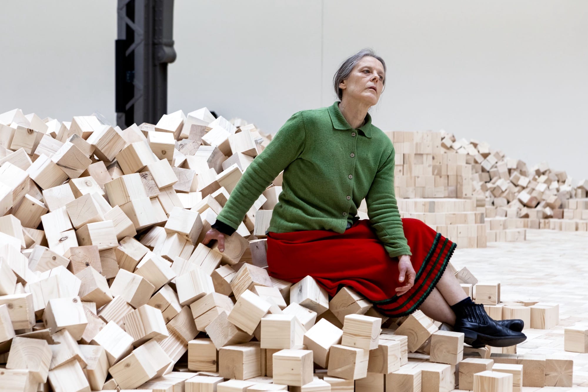 a woman sits on an installation of wooden blocks by Lina Lapelytė in a cavernous building