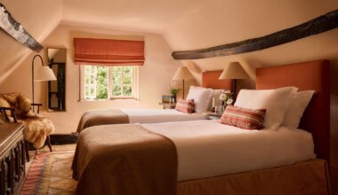 A loft bedroom with sloped ceilings color-drenched plaster pink, an exposed wood ceiling beam, and twin beds with terracotta headboards, crisp white bedding and terracotta cushions and throws.