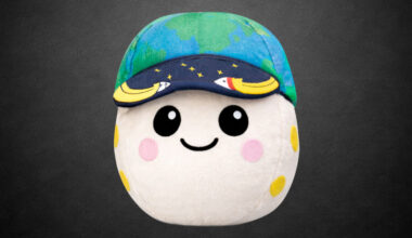 a plush moon with pink and yellow craters, two eyes and a smile, wearing a blue and green cap