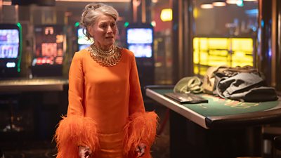 A woman in an orange dress with feathered cuffs stands in a casino looking distressed