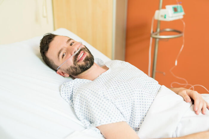 Patient in hospital gown lying in bed with oxygen tube smiling, related to surgical staff revealing patient confessions under sedation.
