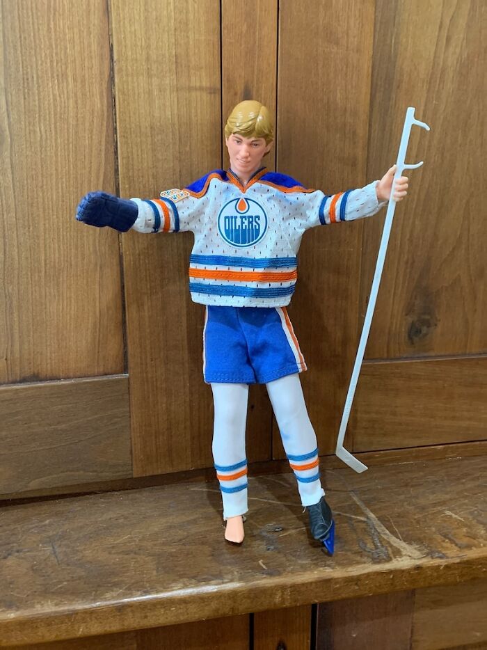 Action figure wearing Edmonton Oilers hockey jersey and gear, standing on wooden surface with a hockey stick.