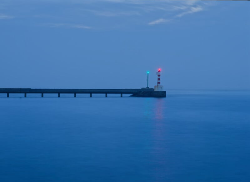 A lighthouse with green and red lights stands at the end of a long pier, surrounded by calm blue water and under an overcast sky at dusk.