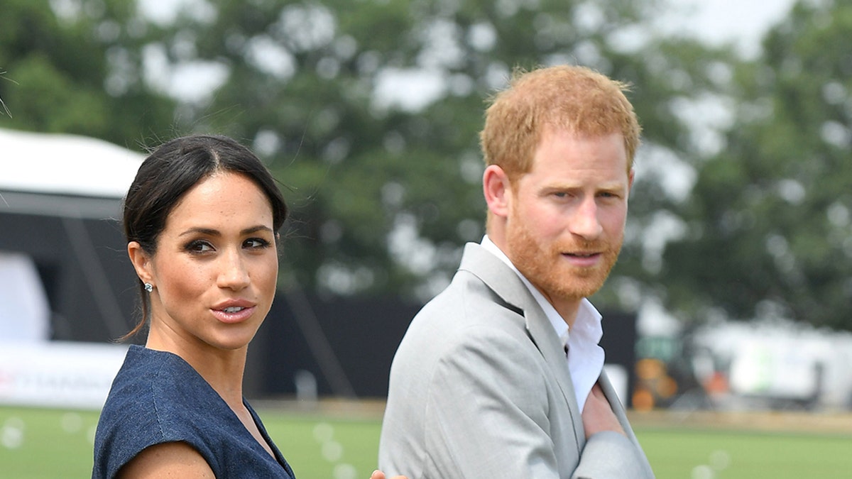 Meghan, Duchess of Sussex, wearing a blue dress, stands next to Prince Harry, Duke of Sussex, in a khaki coat at a polo event