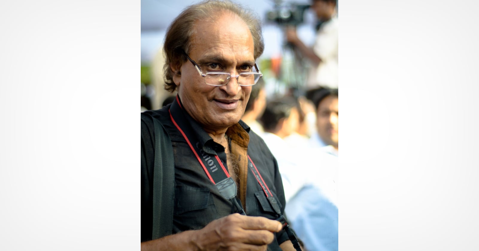 An older man with glasses holding a camera looks at the viewer and smiles. He is wearing a black shirt and a camera strap around his neck, with blurred people and outdoor activity in the background.