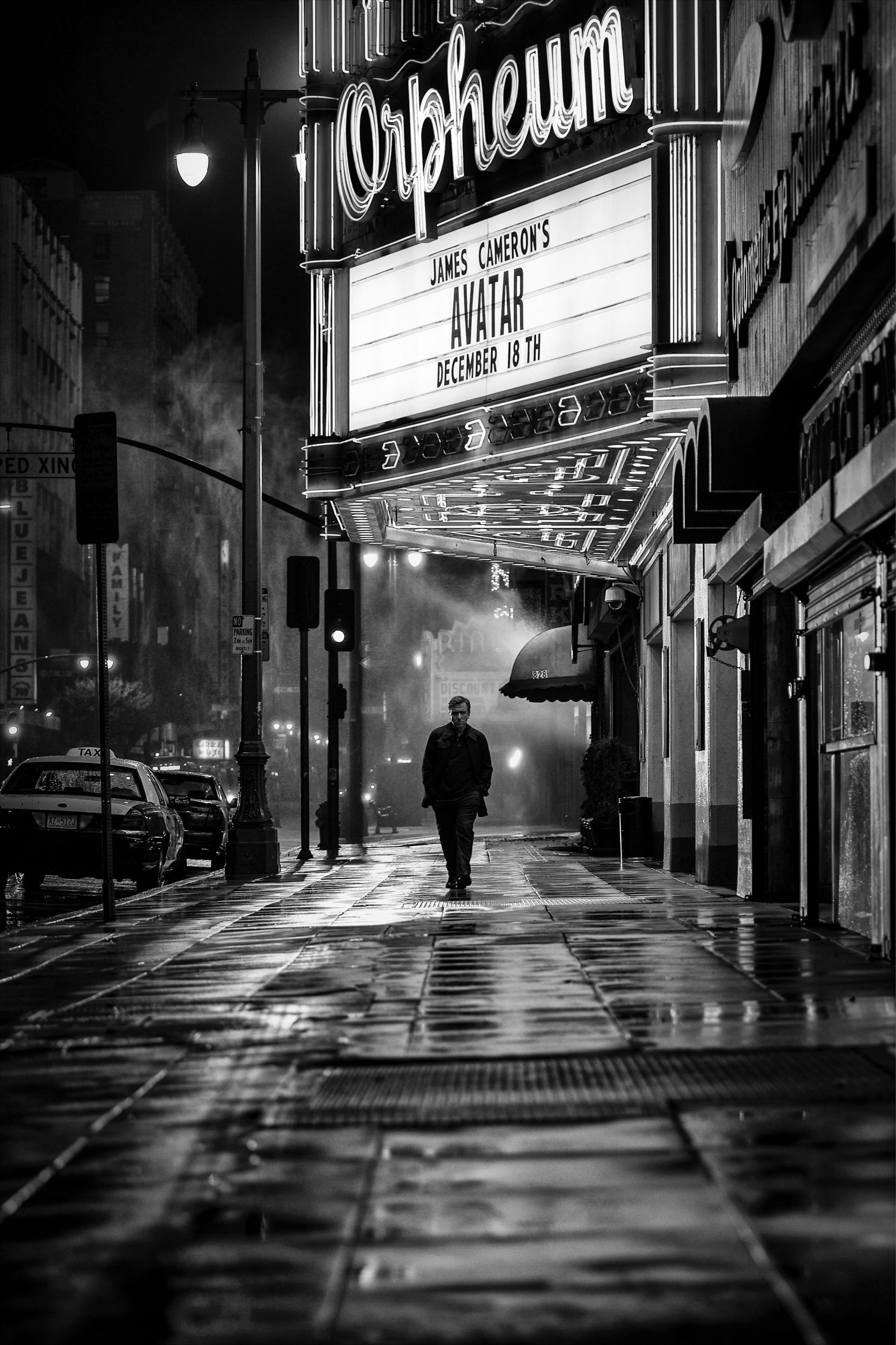 a black-and-white photograph by Daniel Sackheim in a noir style of a man walking down a rainy street below a theater marquis