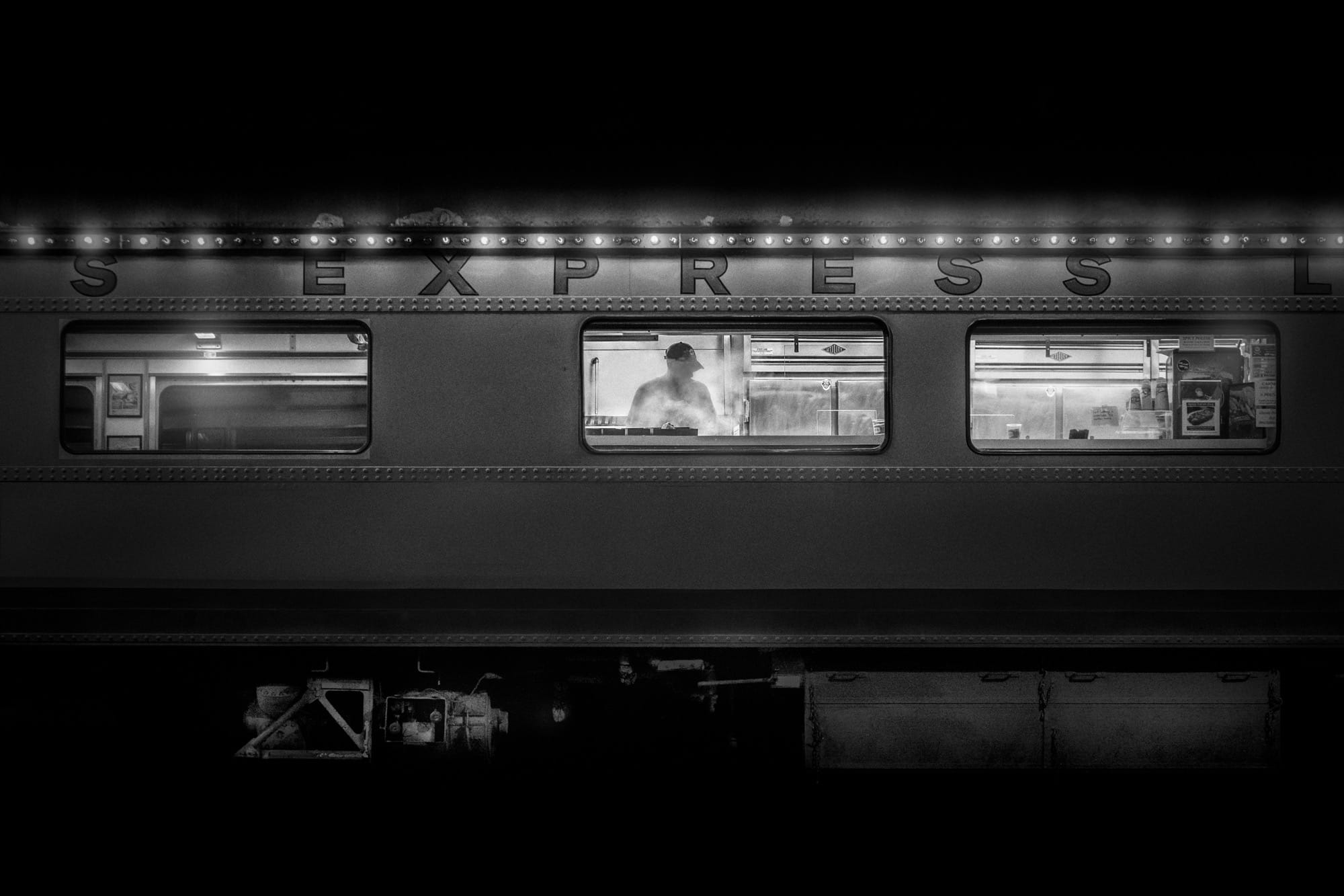 a black-and-white photograph by Daniel Sackheim in a noir style of a cook working in a train car