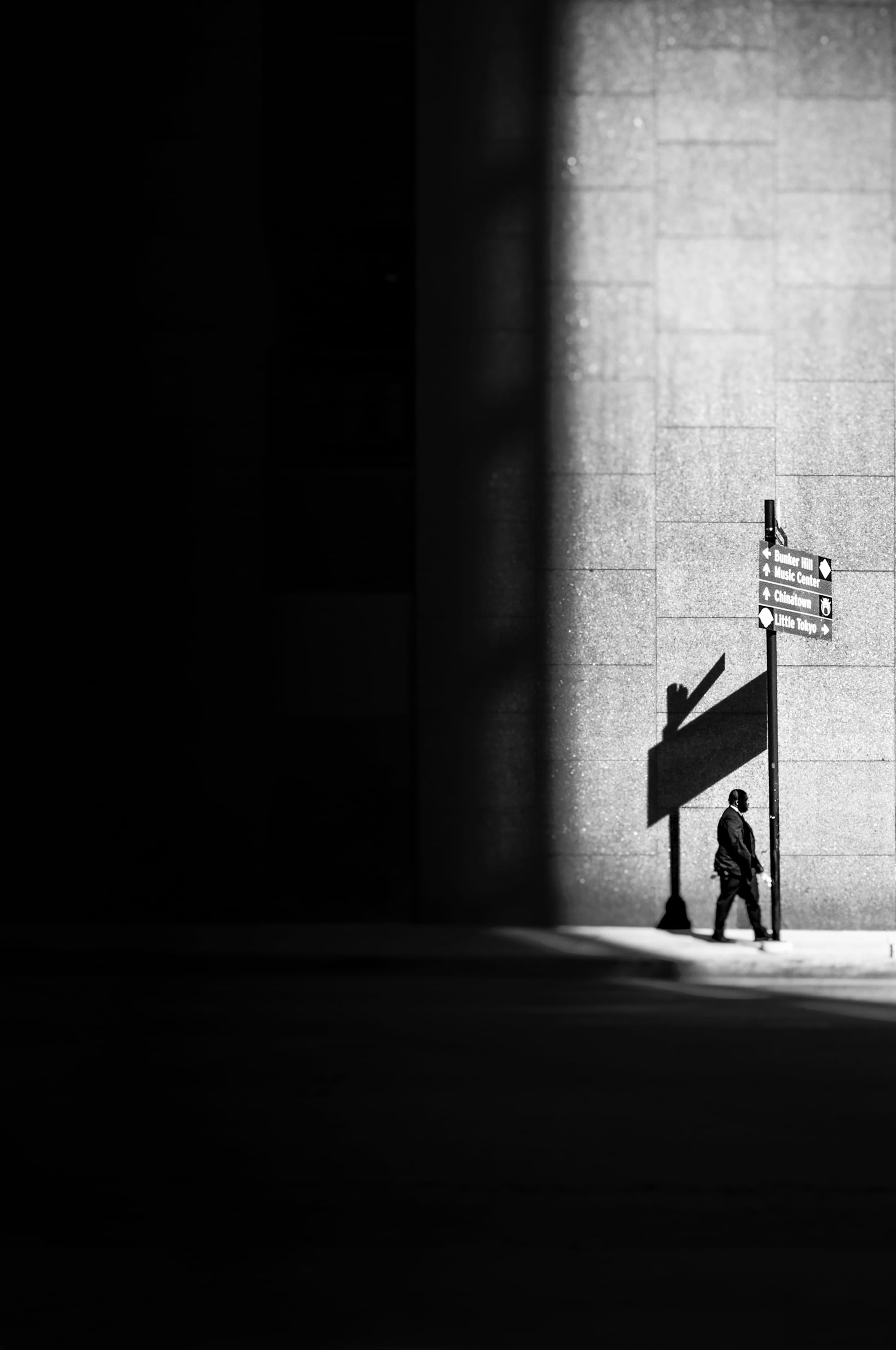 a black-and-white photograph by Daniel Sackheim in a noir style of a figure walking next to a large building amid deep shadows