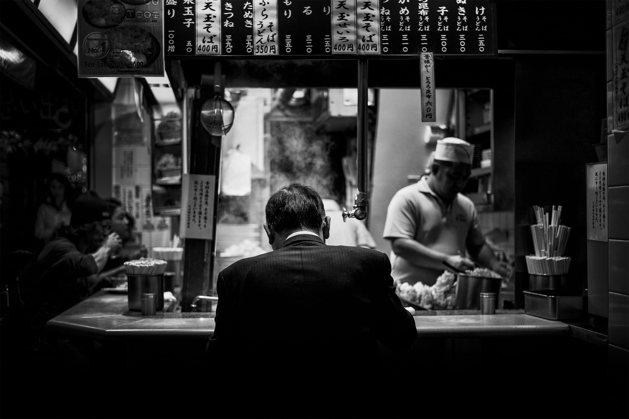 a black-and-white photograph by Daniel Sackheim in a noir style of a man eating at a Japanese food counter