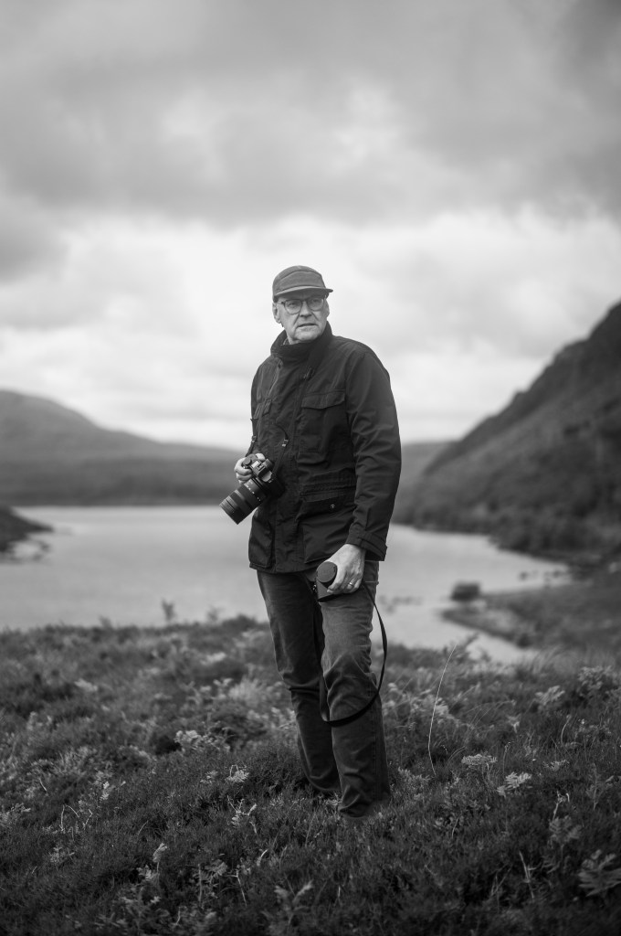 Scott Perkins standing in a rugged lakeside landscape with a camera, photographed in black and white.