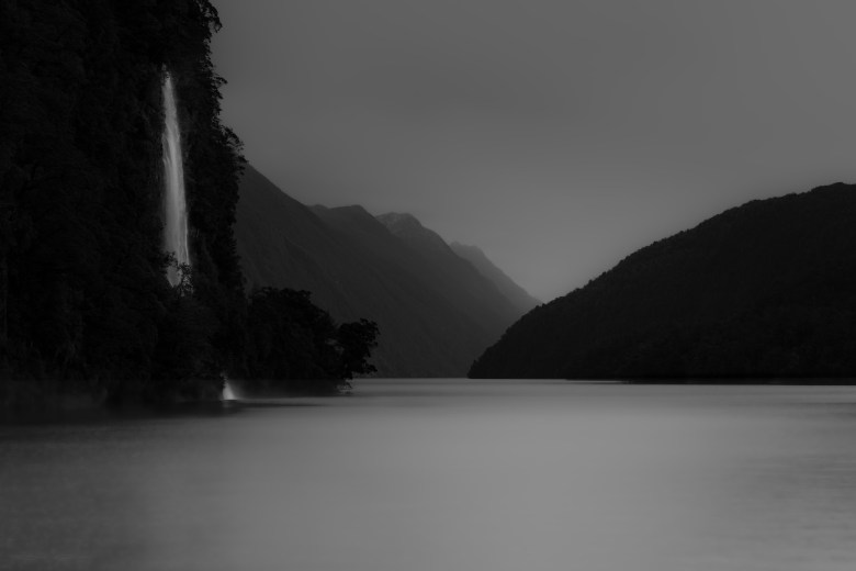 Black-and-white landscape photograph of a waterfall descending into a dark fiord by Scott Perkins.