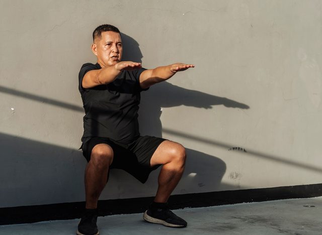 Middle-aged Southeast Asian man holds a wall sit with arms raised, showing control, endurance and core activation during bodyweight isometric training.