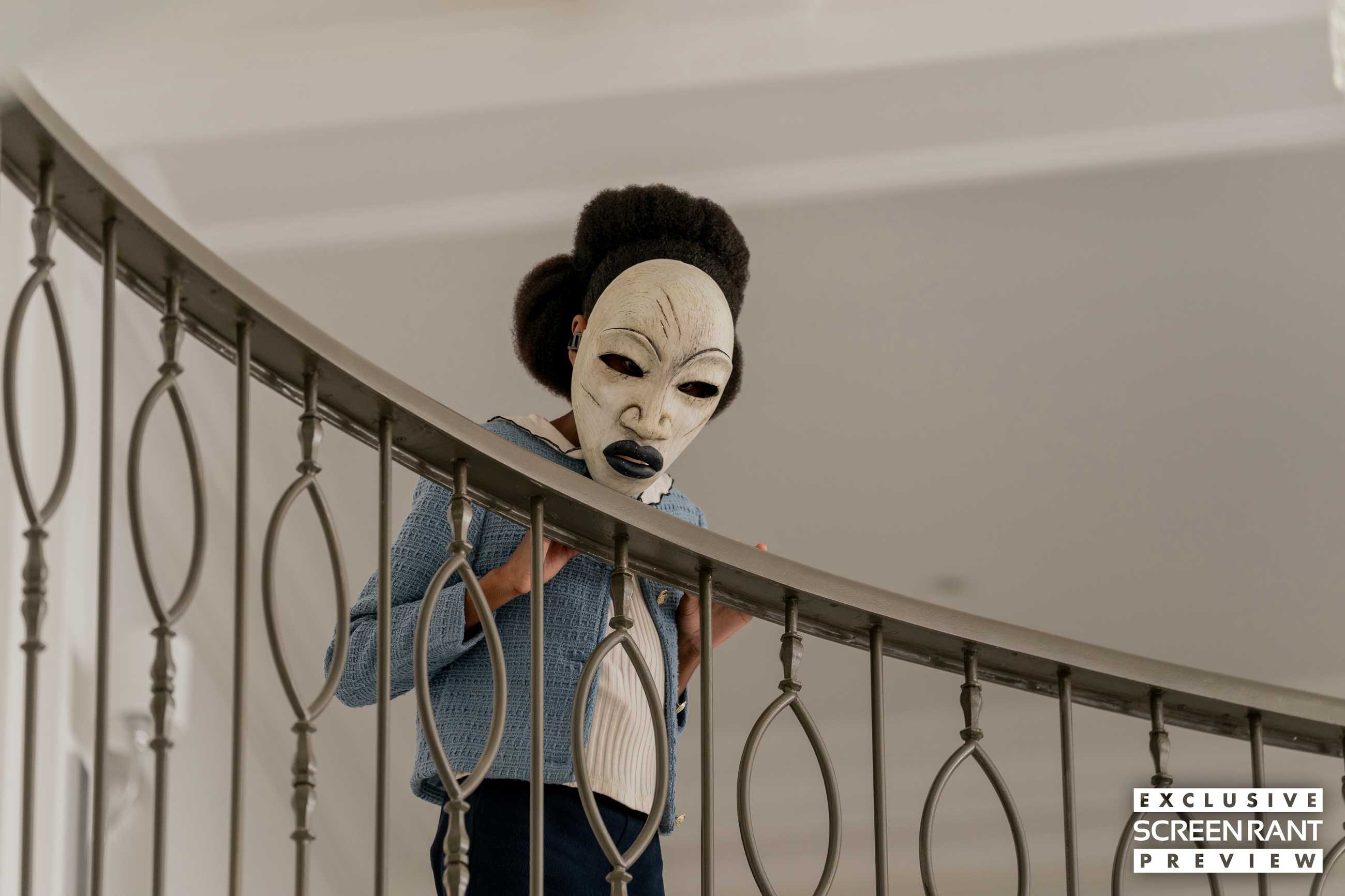 A girl wearing a white mask with black lips standing against a railing in Strung
