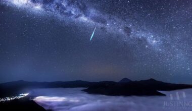 The Milky Way stretches across the sky and a bright Eta Aquarid meteor streaks across the center. Below is the top of Mount Bromo surrounded by clouds.