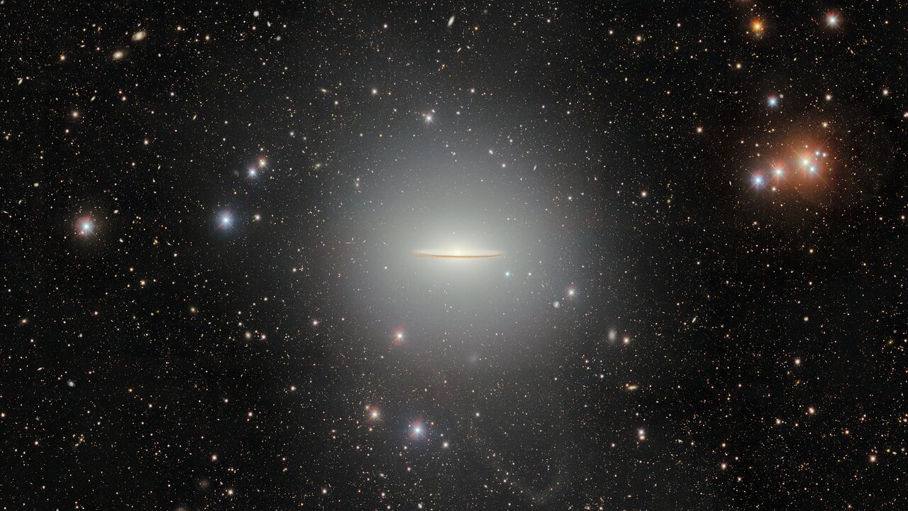 A black background of space is speckled with a wide array of stars. At the center is a flat, glowing galaxy shaped like a stretched out sombrero.