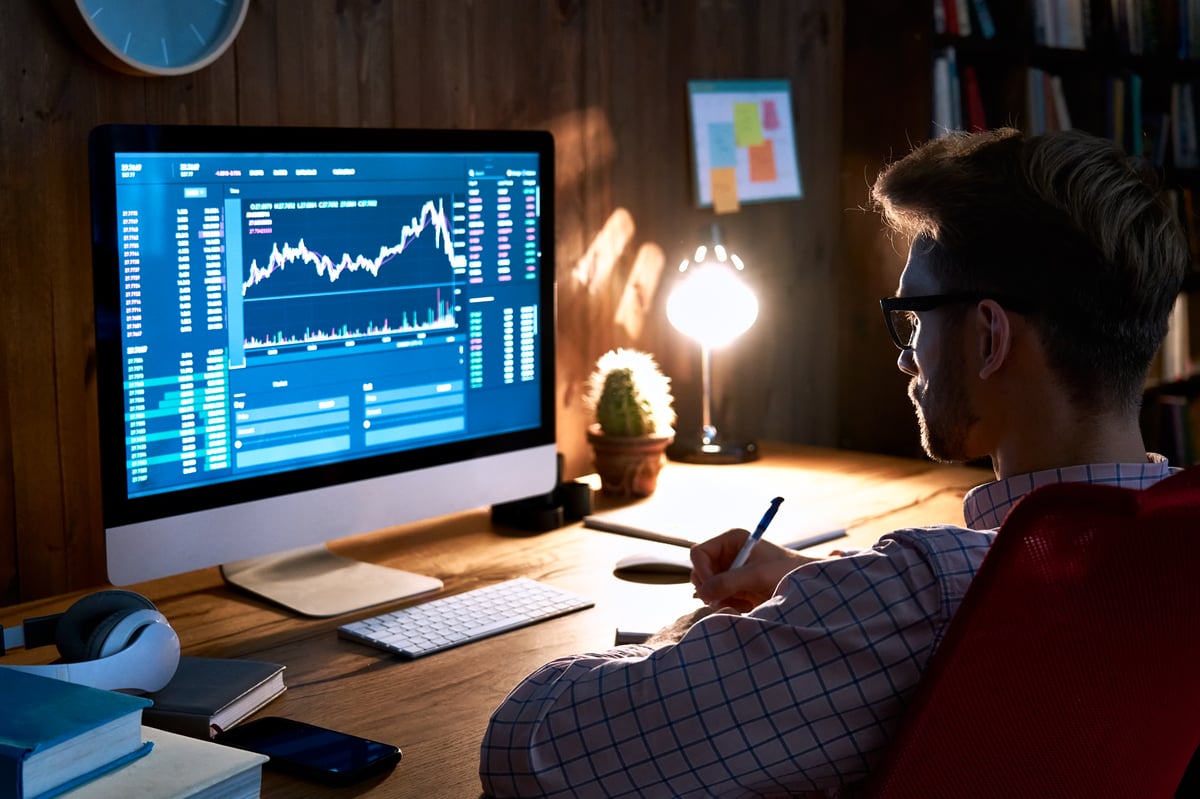 An investor sitting in a dark room looking at their computer screen, which is displaying a stock or cryptocurrency chart.