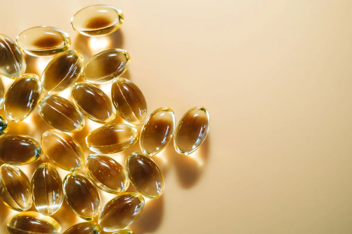 Softgels with transparent yellow oily substance reflecting sunlight on beige background.