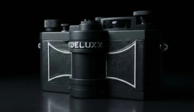 A black vintage-style camera labeled “DELUX” is shown against a dark background, with textured panels and metallic knobs visible in dramatic lighting.