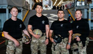 four young military men wearing black t-shirts and camouflage pants stand on the deck of a ship at sea