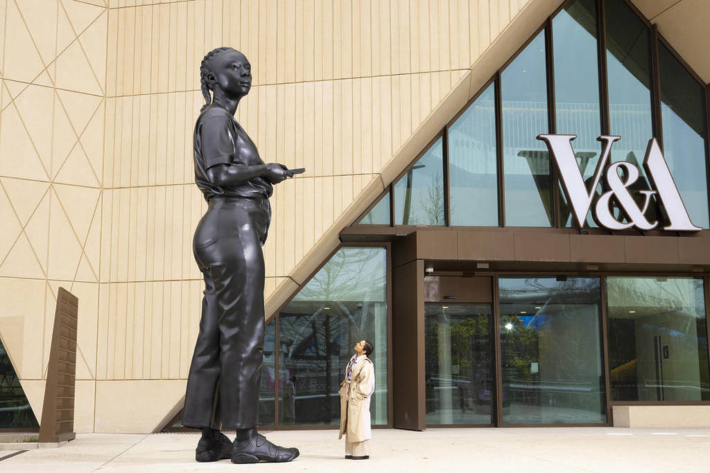 someone standing outside the entrance to V&A East gazing up at an 18ft tall statue of a person