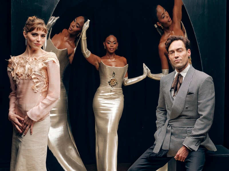 Three people pose stylishly: a woman in a pale pink dress with feather details, a woman in a shimmering silver gown with long gloves, and a man in a patterned suit. Their reflections appear in a glossy black arch behind them.