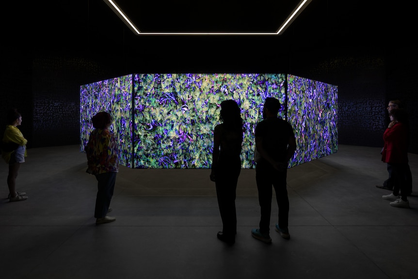 A group of people look at a series of screens hung in the air. The screens show intricate purple and blue patterns pulsating.