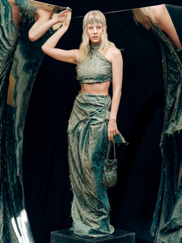 A woman in a shiny sage green two-piece outfit stands on a pedestal, holding a small matching bag. She wears a jeweled headpiece and poses against curved, reflective metallic panels.