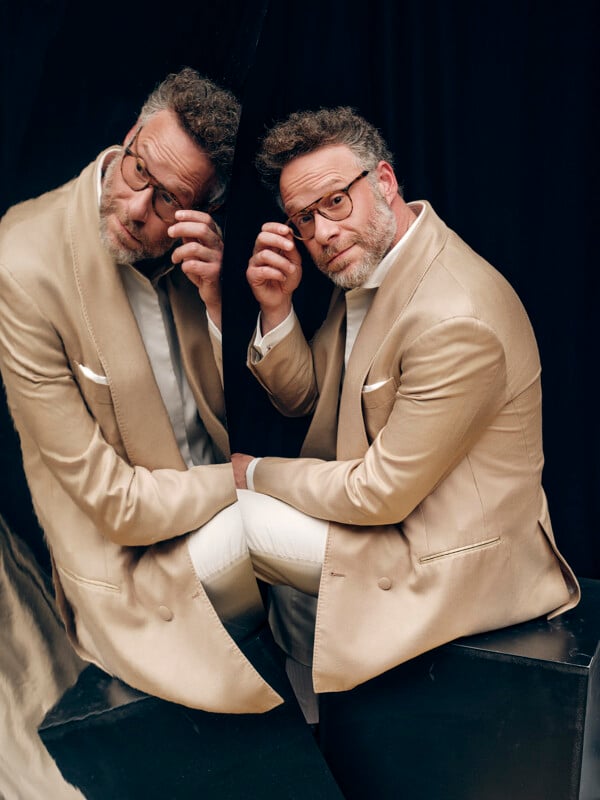 A man in a beige suit and glasses sits on a black platform, touching his glasses and looking into a mirror, creating a reflection of himself against a black background.