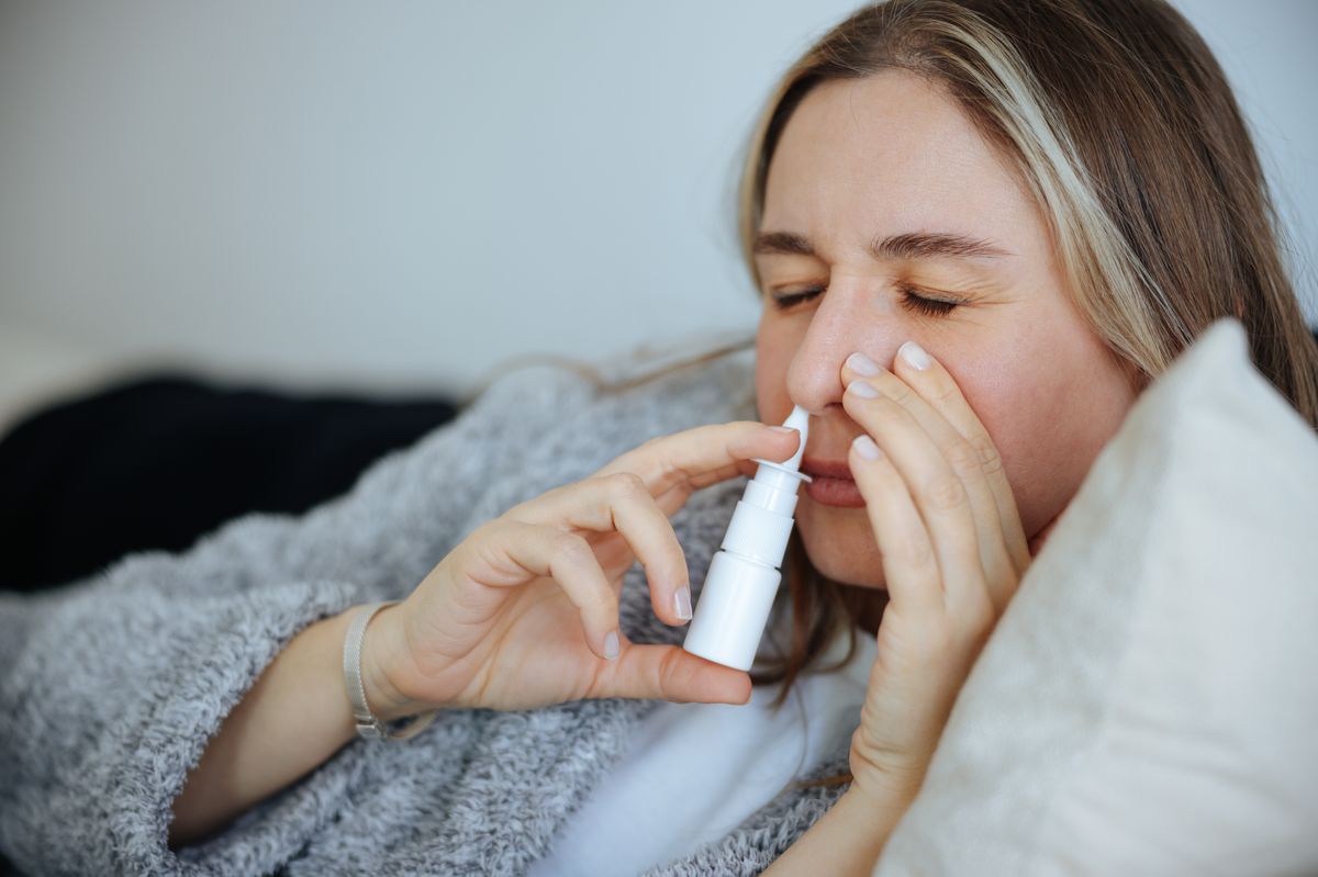 Woman is shown using a nasal spray in her bedroom to alleviate her congestion. Treatment for Allergies or Common Cold. Woman Using Nasal Spray Drops of Medicine in Her Nose. Rhinitis, Allergy.