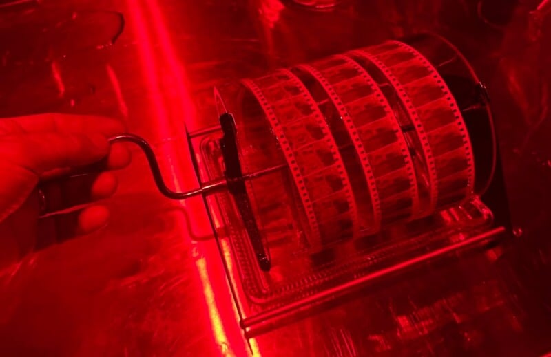 A hand turns a film reel crank under red darkroom lighting. Several loops of photographic film are wound around the reel, which is placed on a reflective metal surface.