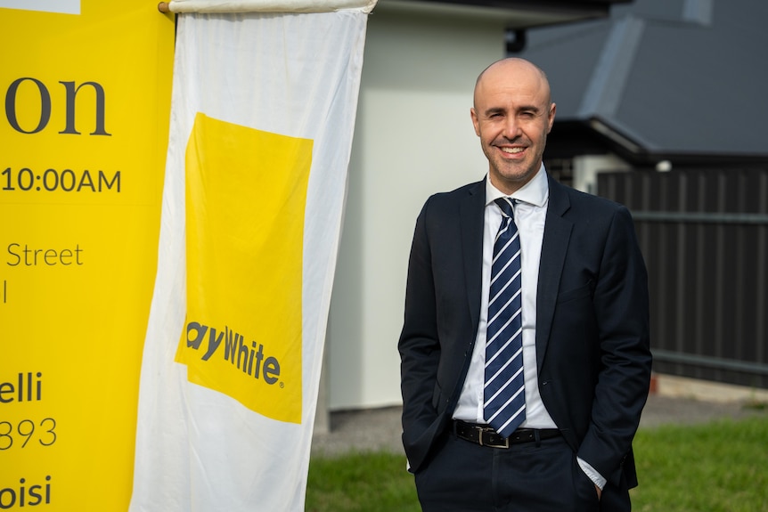 A man wearing a suit and tie stands next to a Ray White flag with his hands in his pockets, smiling. 