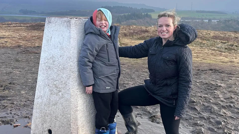Arthur, aged 8, wearing a grey coat and a turqoise hoody, smiles at the camera on a hilltop next to his mum, Jessica Pryce, who has wavy blonde hair tied back and is wearing a blue coat.