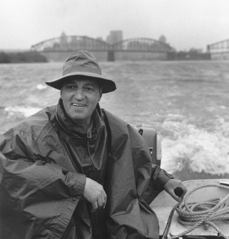 A man wearing a rain hat and poncho sits in a small boat on a river, smiling. The water is choppy, and a bridge with arched supports is visible in the background on a cloudy day.