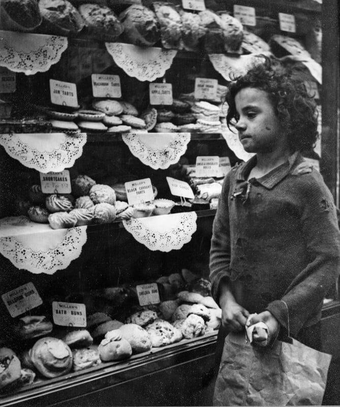 A young girl with curly hair stands by a bakery window displaying various pastries and cakes, holding a paper bag and a bun. The shelves are lined with doilies and labeled signs for items like jam tarts and Bath buns.