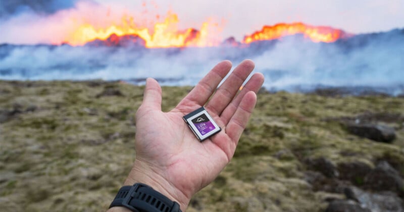 A hand holding an SD card in the foreground, with a rocky, grassy terrain and a large fire or lava eruption in the blurred background.