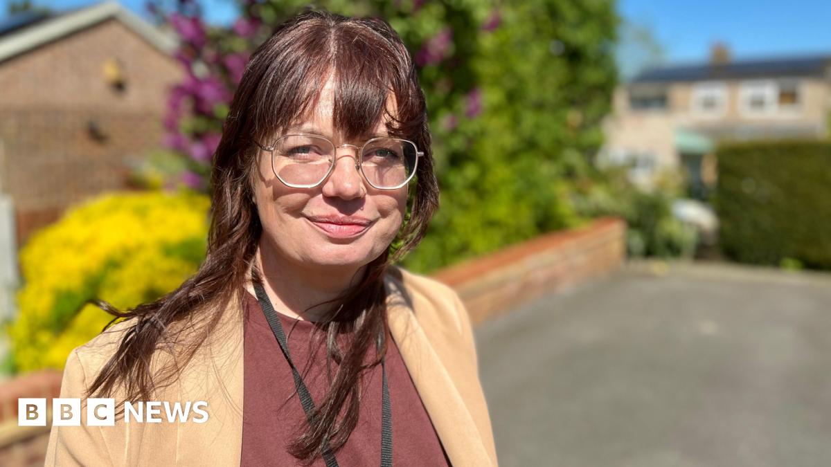Claire Colvill, a social work apprentice, looks at the camera outside a client's home in Hertfordshire. She is wearing a light brown suit jacket with a maroon smart T-shirt underneath it.  She has a black lanyard around her neck and is wearing metal framed glasses. She has brown hair with a fringe that has a red tone. She is standing in front of a driveway with a brick wall and hedges along the wall.