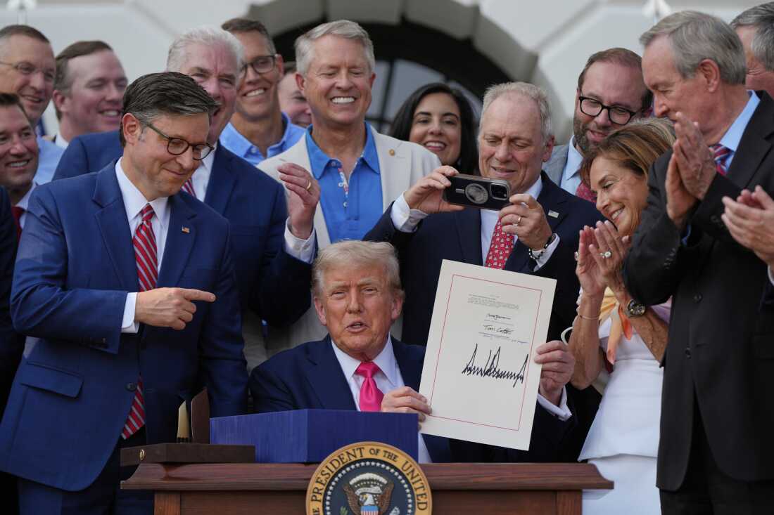 President Donald Trump is shown proudly holding up a piece of paper that shows his distinctive large, Sharpie-penned signature. He is surrounded by many other men and one woman.