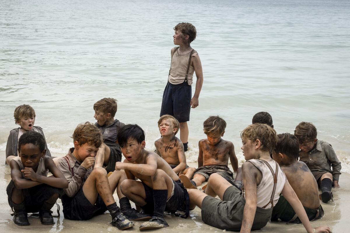 A group of young, dirty children sit on the beach as waves crash into them. One boy is standing.