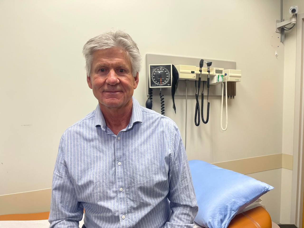 A man with grey hair is wearing a blue and white striped dress shirt. He's sitting on an exam bed. Medical equipment including a blood pressure cuff is hanging on the wall behind him