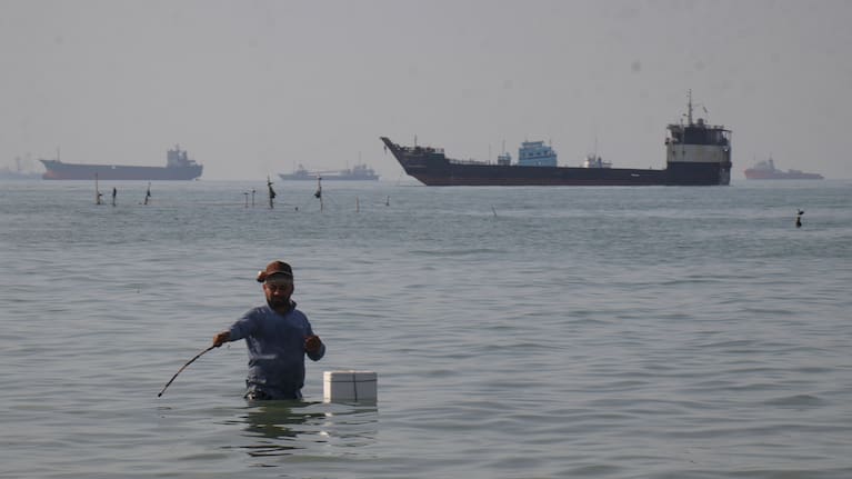 A man stands in the water, appearing to fish, as bulk carriers, cargo ships, and service vessels line the horizon in the Strait of Hormuz off Bandar Abbas, Iran