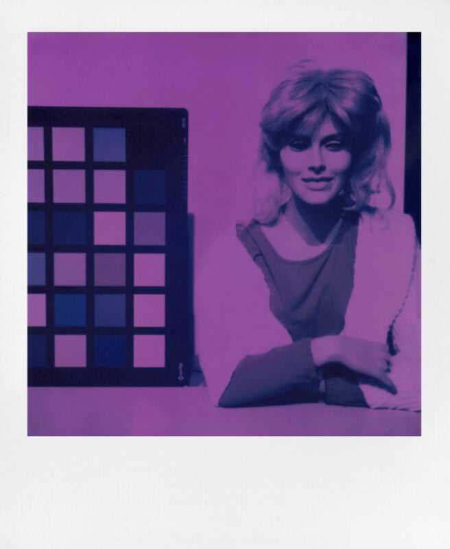 A woman with shoulder-length hair sits at a table next to a color calibration chart. The image is tinted purple, giving it a surreal effect. The woman is smiling gently and resting her arms on the table.