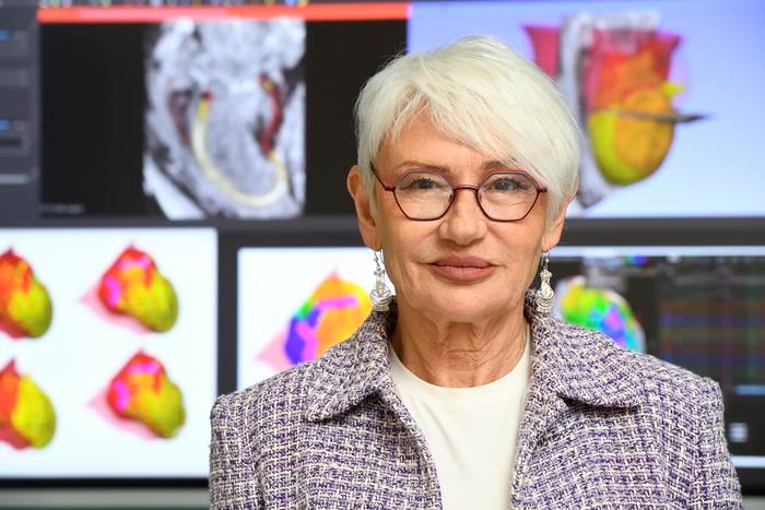 A striking older woman with short white hair looks into the camera as she stands in front of colourful images of the virtual hearts.