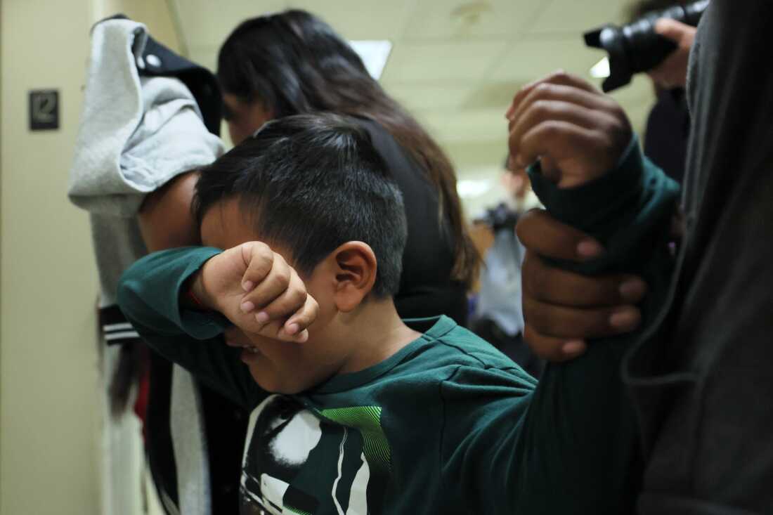 A child who appears to be about 8-years-old holds his arm over his eyes as he cries. An adult his holding his other arm. 