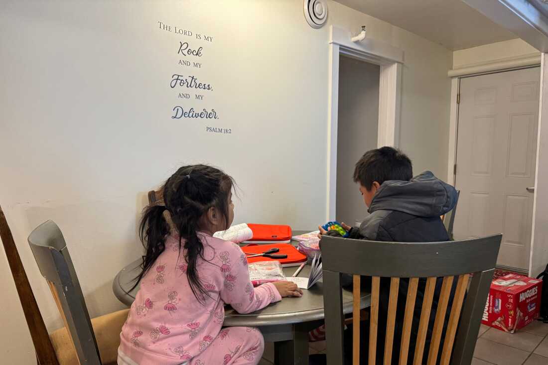 Two children, who appear to be a 7-year-old girl and a 9-year-old boy, sit at a kitchen table with coloring books. A Bible verse is on the wall above them. It reads, "The Lord is my rock and my fortress and my deliverer -- Psalms 18:2." 